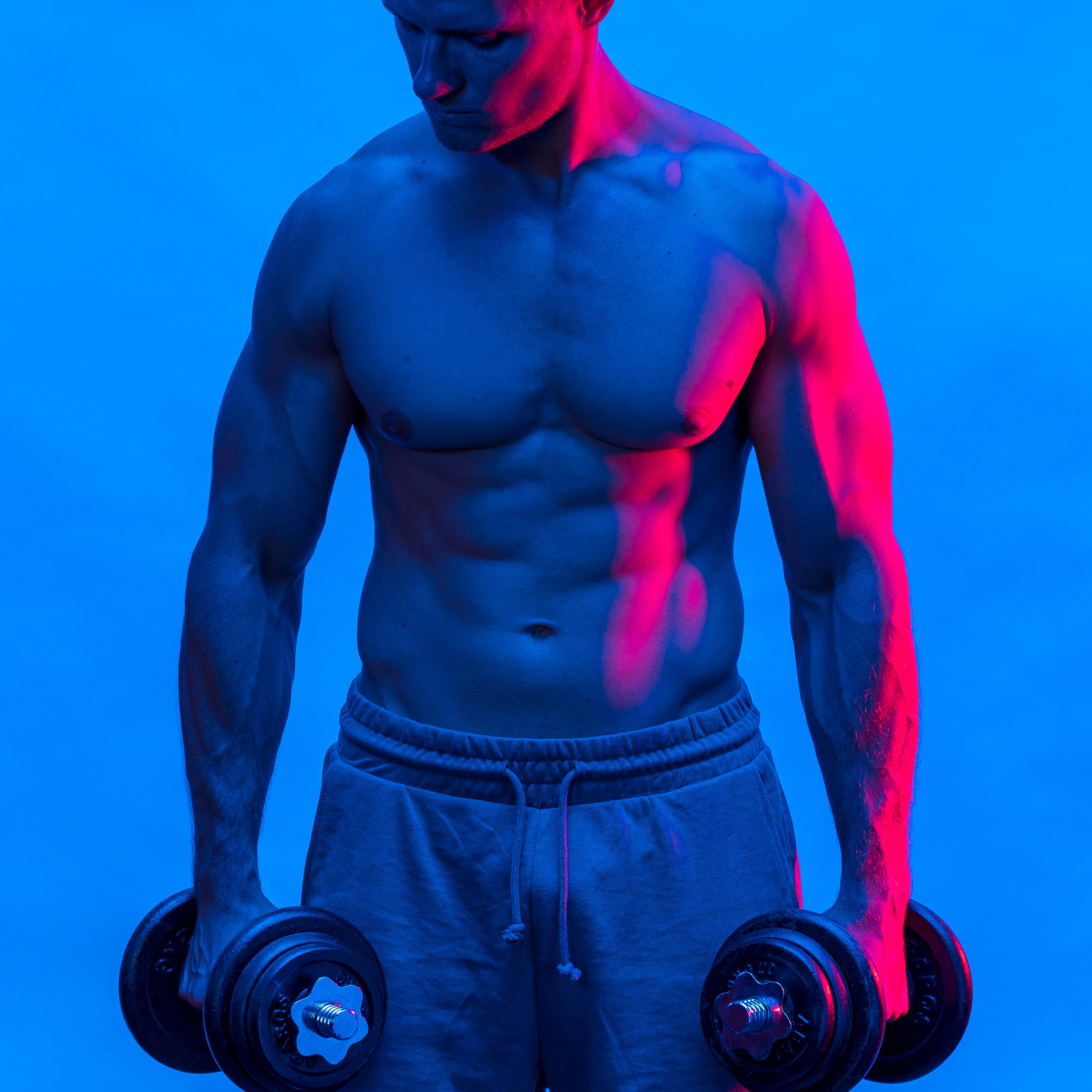 Shirtless man with a muscular build, holding a dumbbell in each hand, showcases testosterone enhancement and overall male hormonal health.