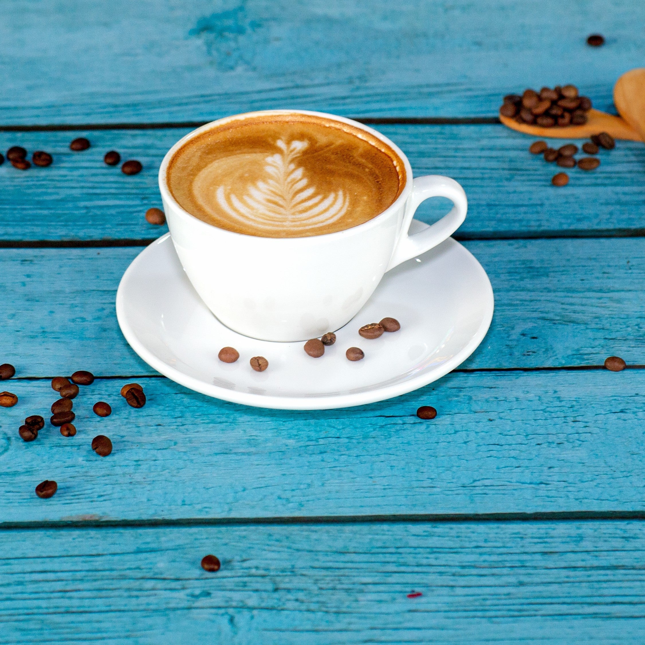 White coffee cup filled with latte and leaf-shaped latte art on saucer surrounded by coffee beans on blue wooden surface, ideal pre-workouts boost and endurance.