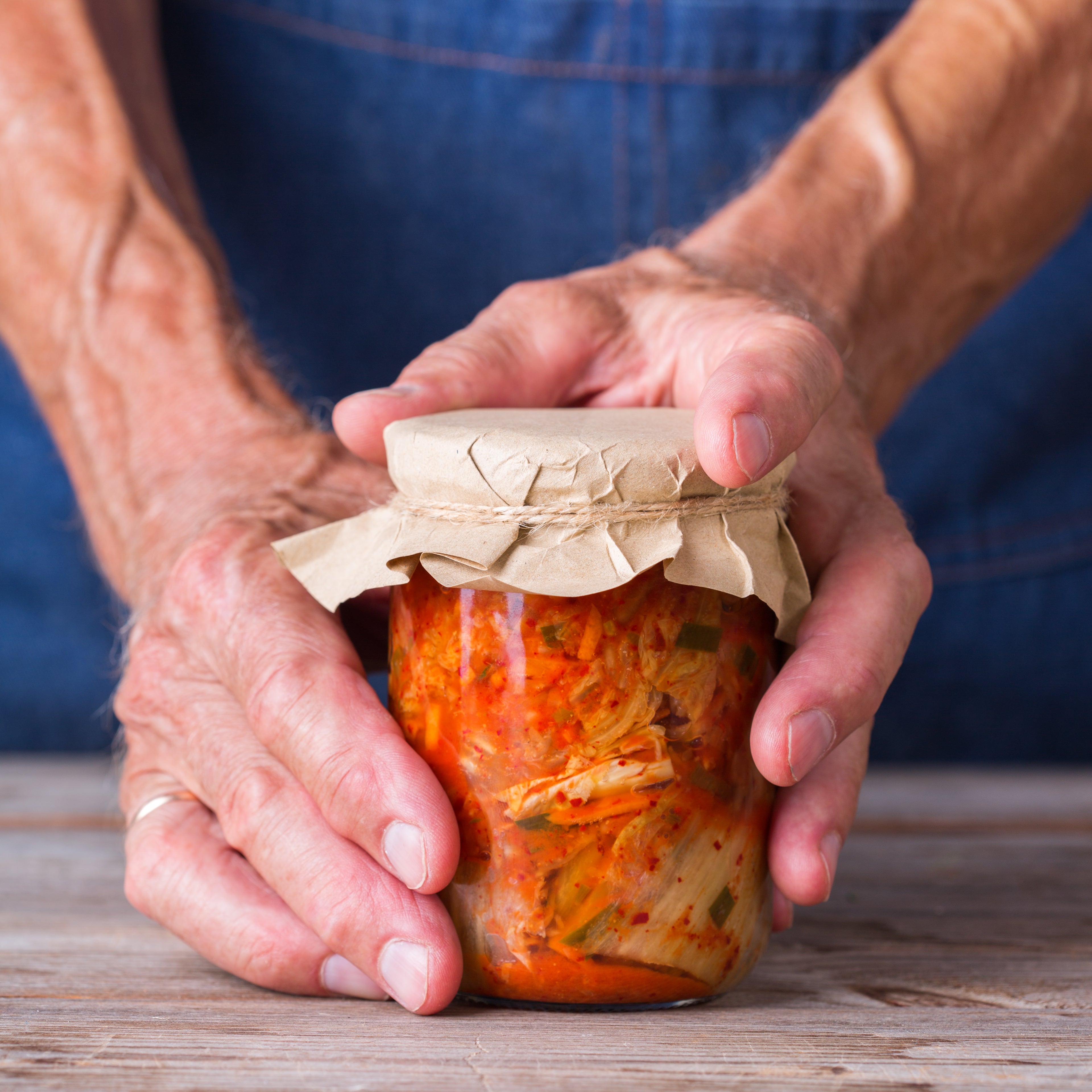 Hands holding a jar of homemade kimchi with a paper cover tied with string on wooden surface, embodying postbiotics for gastrointestinal wellness.