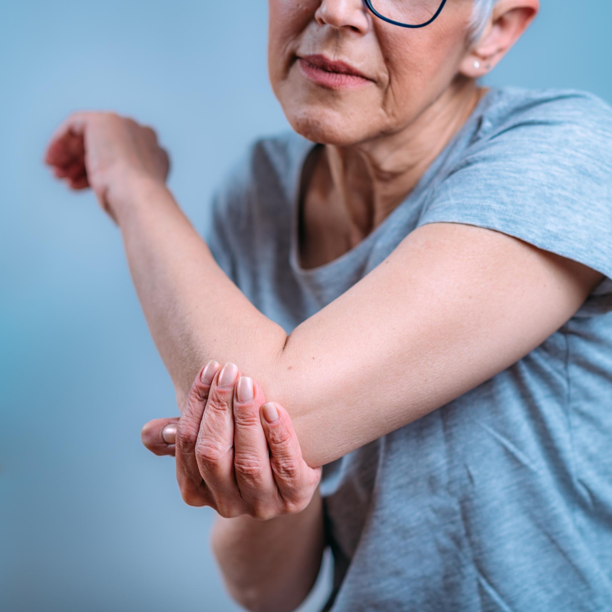 Elderly woman in gray shirt holds elbow appearing to experience discomfort, emphasizing joint support for active, pain-free living.