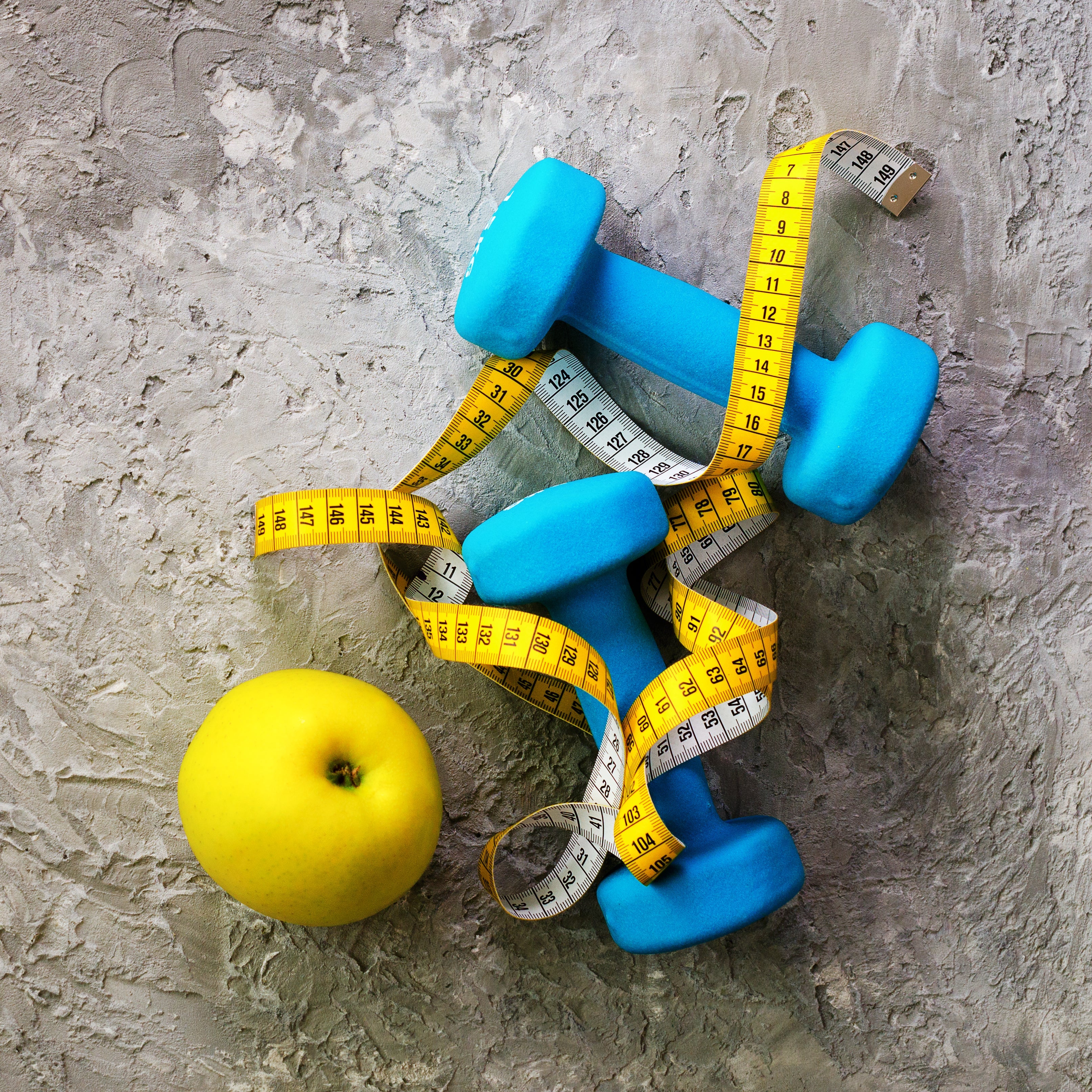 Yellow apple with blue dumbbells, and measuring tapes on gray surface represents weight loss efforts and appetite control strategies.